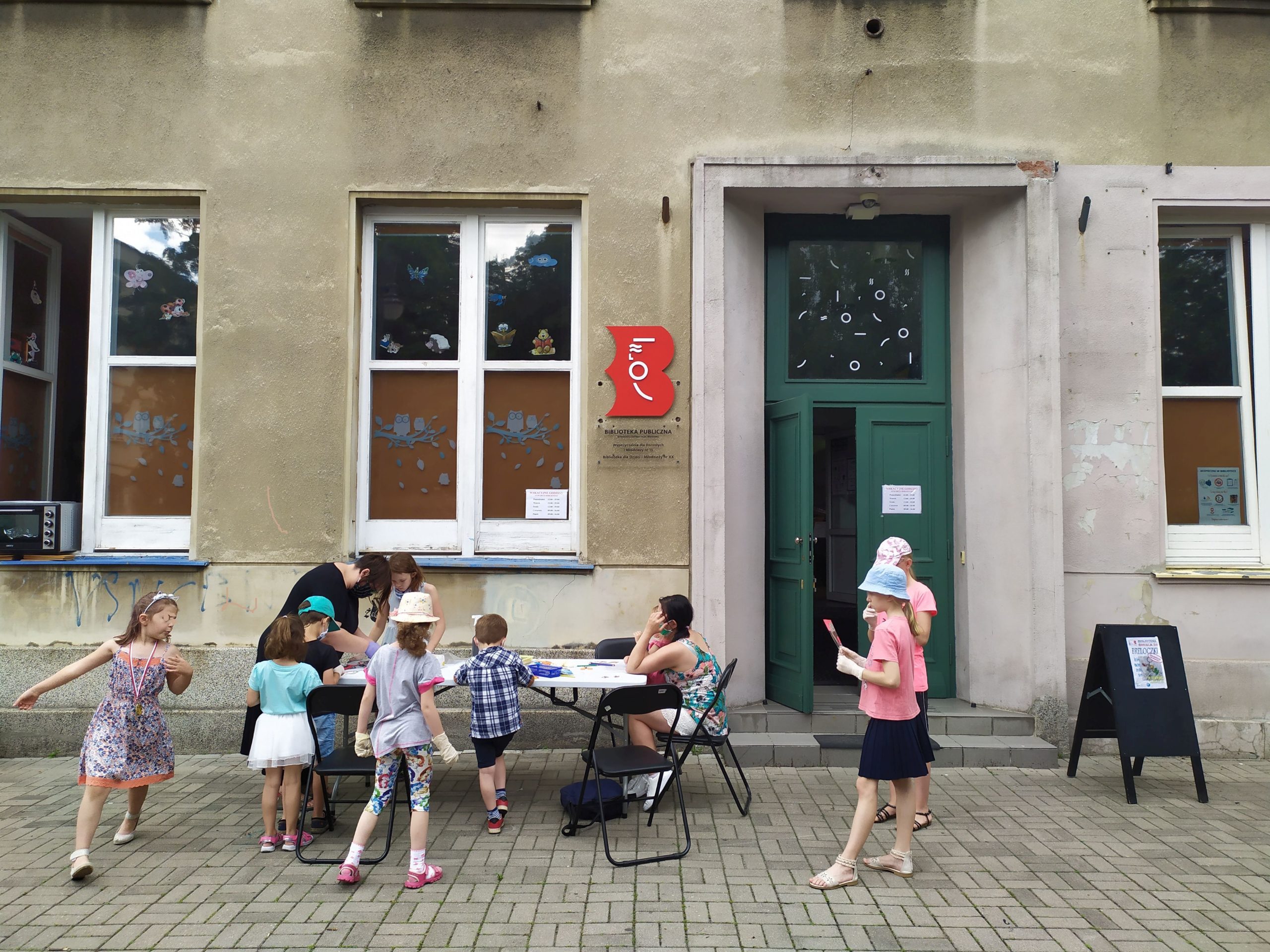 Biblioteka przy ul. Śmiałej 24 widok z zewnątrz na wejście główne. Grupa dzieci bierze udział w warsztatach prowadzonych przed Biblioteką