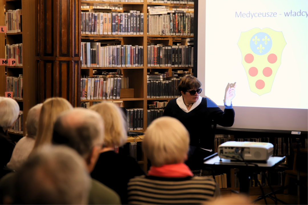 Zdjęcie z trzeciej prelekcji z cyklu wykładów o sztuce i historii: "Medyceusze - władcy Florencji" przedstawiające Panią Magdalenę Tworzowską, slajd na rzutniku z napisem Medyceusze - władcy Florencji oraz słuchaczy podczas wykładu wnętrzu Wypożyczalni nr 16