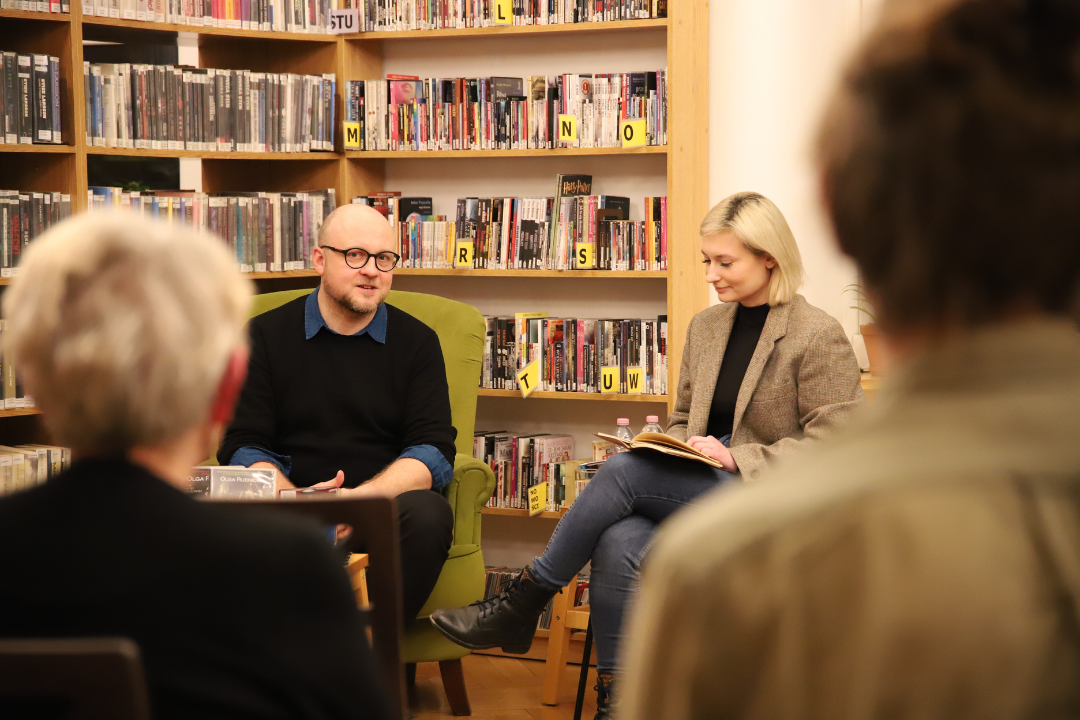 zdjęcie macieja więckowskiego, który siedzi w fotelu oraz siedzącej obok niego aleksandry szatkowskiej a także publiczności na tle regału z audiobookami we wnętrzu wypożyczalni nr 16 podczas spotkania o audiobookach