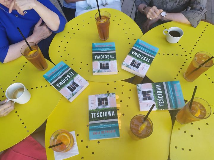 Spotkanie członków Dyskusyjnego Klubu Książki. Klubowicze siedzą przy żółtych złączonych stolikach, na których rozłożone sa książki oraz stoja napoje. 