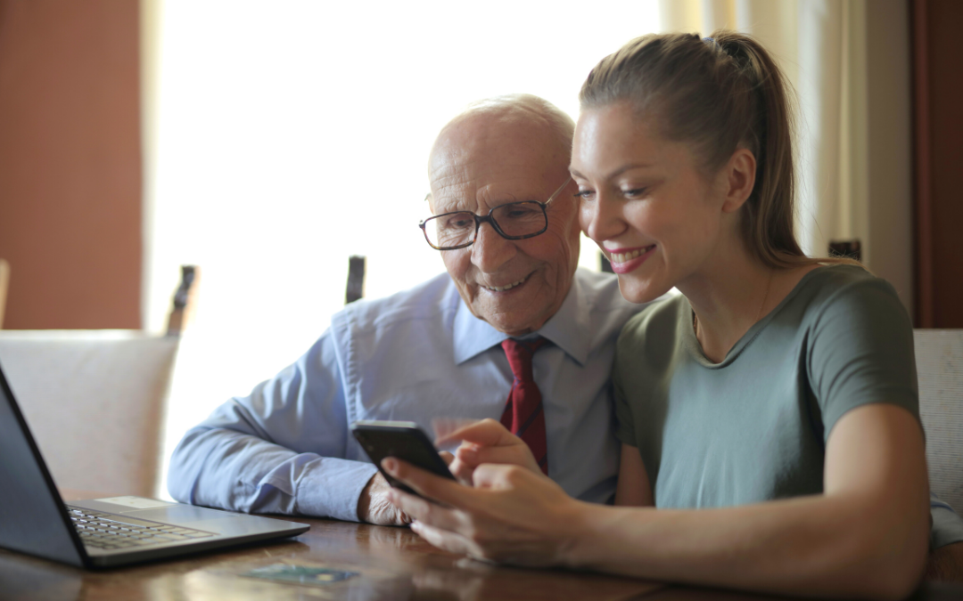 zdjęcie przedstawiające osobę starszą korzystająca ze smartfona oraz osobę młodszą pomagającą w jego obsłudze, z lewej strony zdjęcia jest komputer