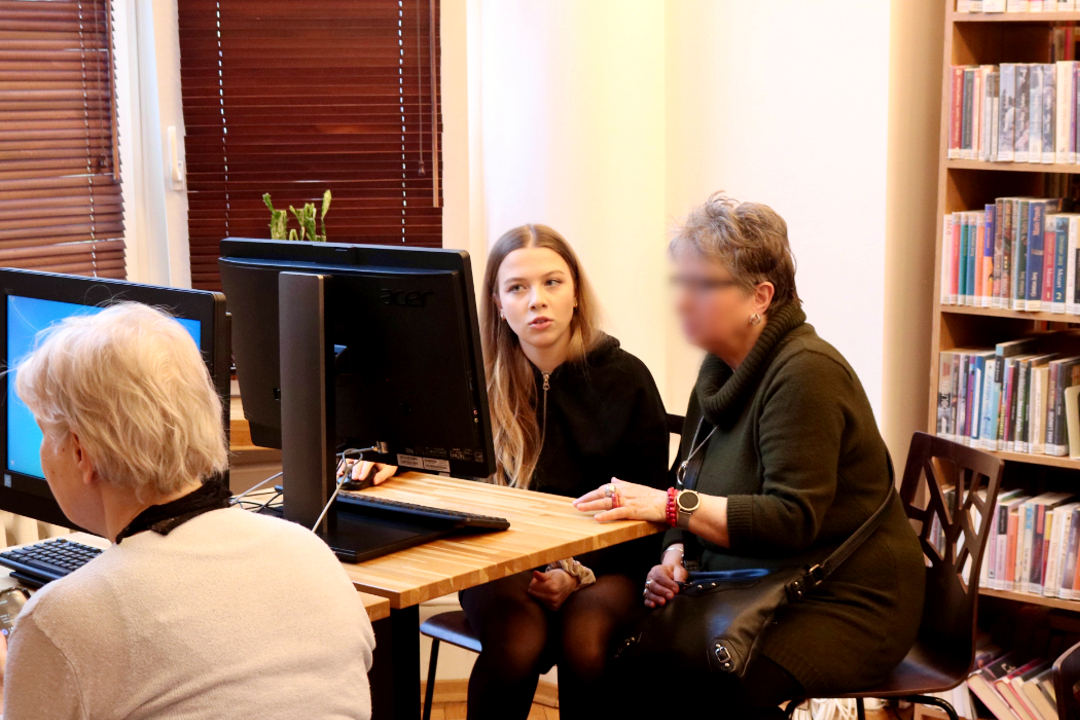 zdjęcie przedstawia wolontariuszkę przy komputerze udzielającą pomocy seniorce przy konkretnym problemie podczas szkolenia Senior bezpieczny w sieci we wnętrzu Wypożyczalni nr 16 (Słowackiego 19a)