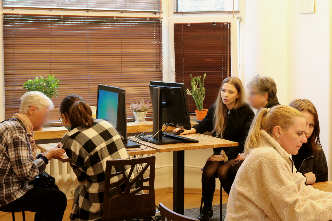 zdjęcie przedstawia wolontariuszy przy komputerach lub urządzeniach mobilnych udzielających pomocy seniorce przy konkretnym problemie podczas szkolenia Senior bezpieczny w sieci we wnętrzu Wypożyczalni nr 16 (Słowackiego 19a)
