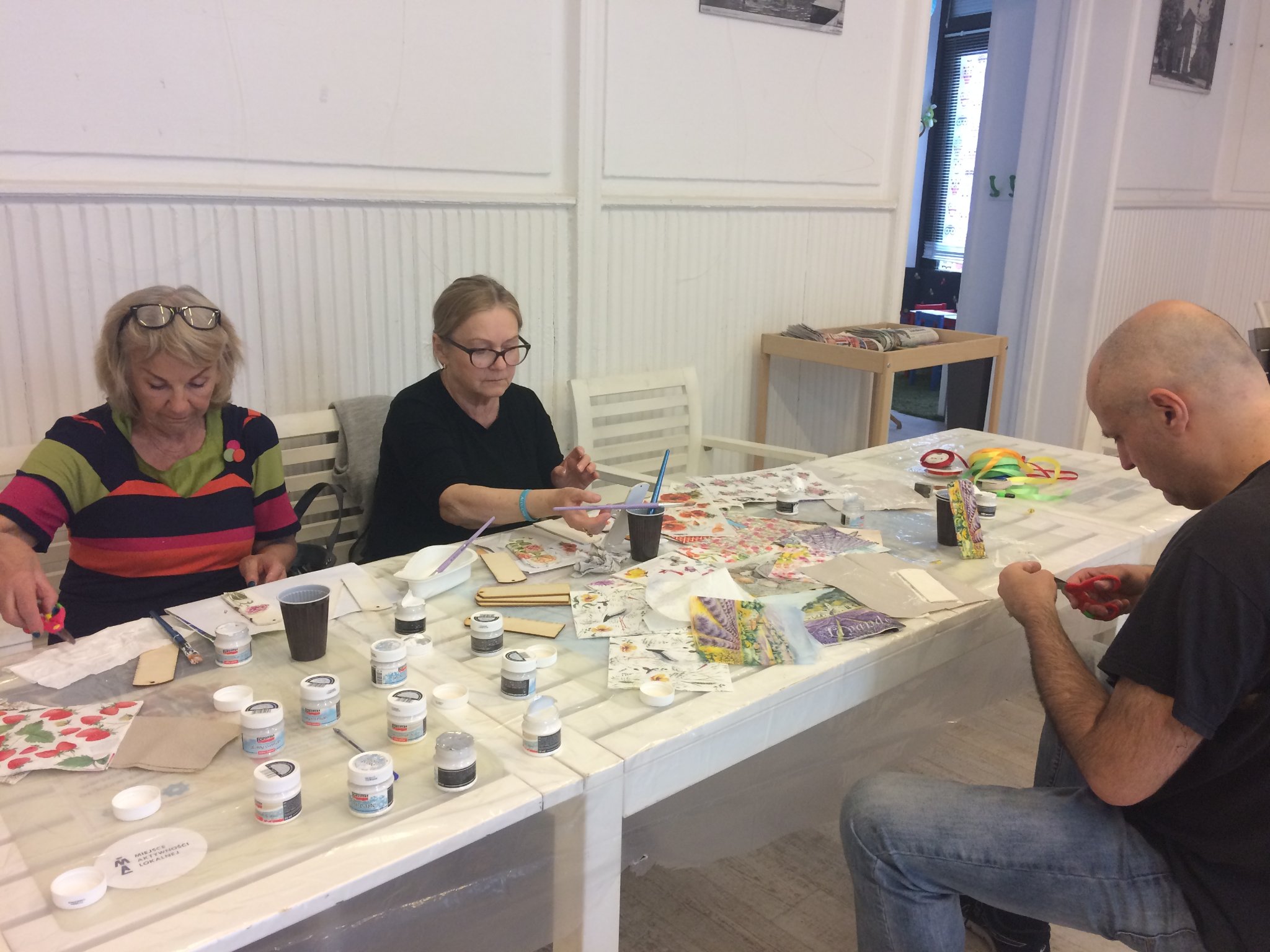 Uczestnicy warsztatów podczas tworzenia zakładek. Na stole rozłożone nożyczki, farby i kolorowy papier
