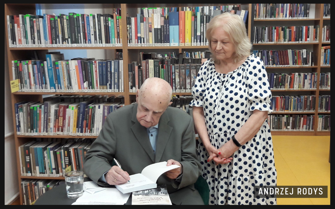 Andrzej Rodys przy stoliku wpisuje dedykacje do książek. Obok niego stoi czytelniczka