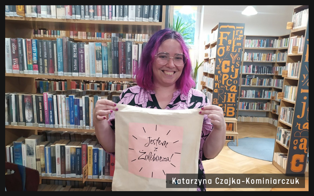 autorka pozuje do zdjęcia na tle bibliotecznych regałów, w rękach trzyma torbę z napisem "jestem z żoliborza"