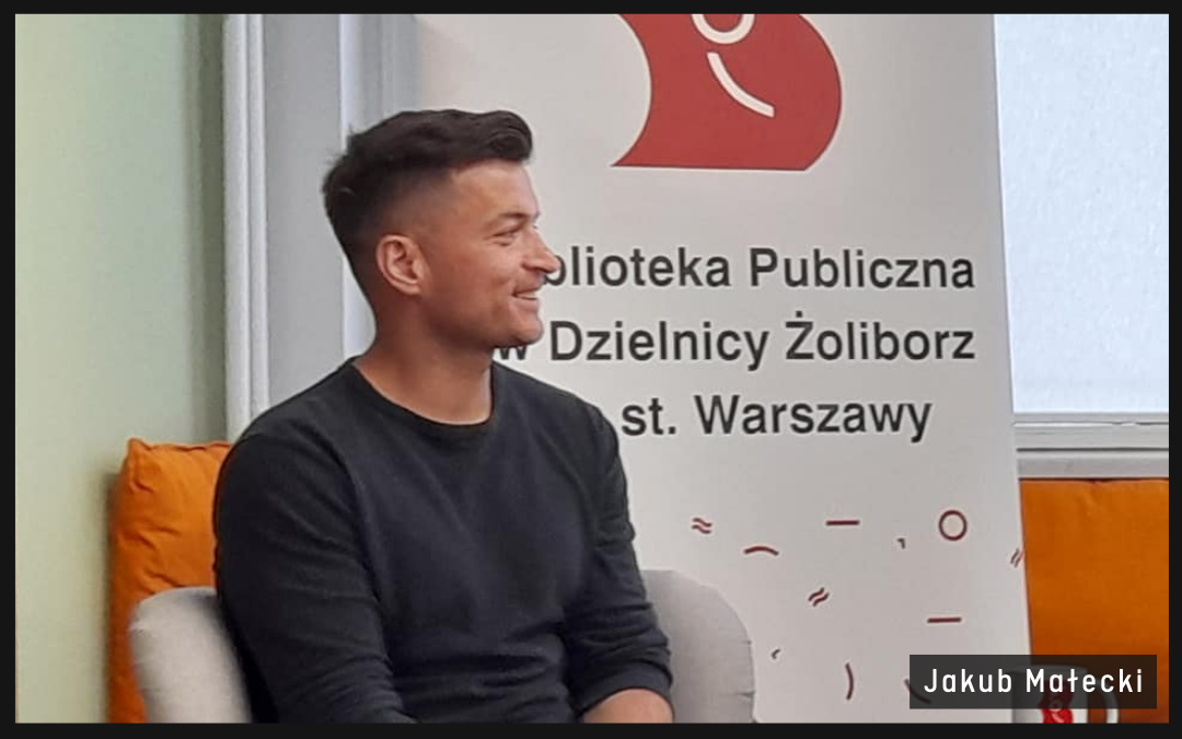 autor siedzi na tle banneru reklamowego biblioteki, w tle czerwone logo biblioteki