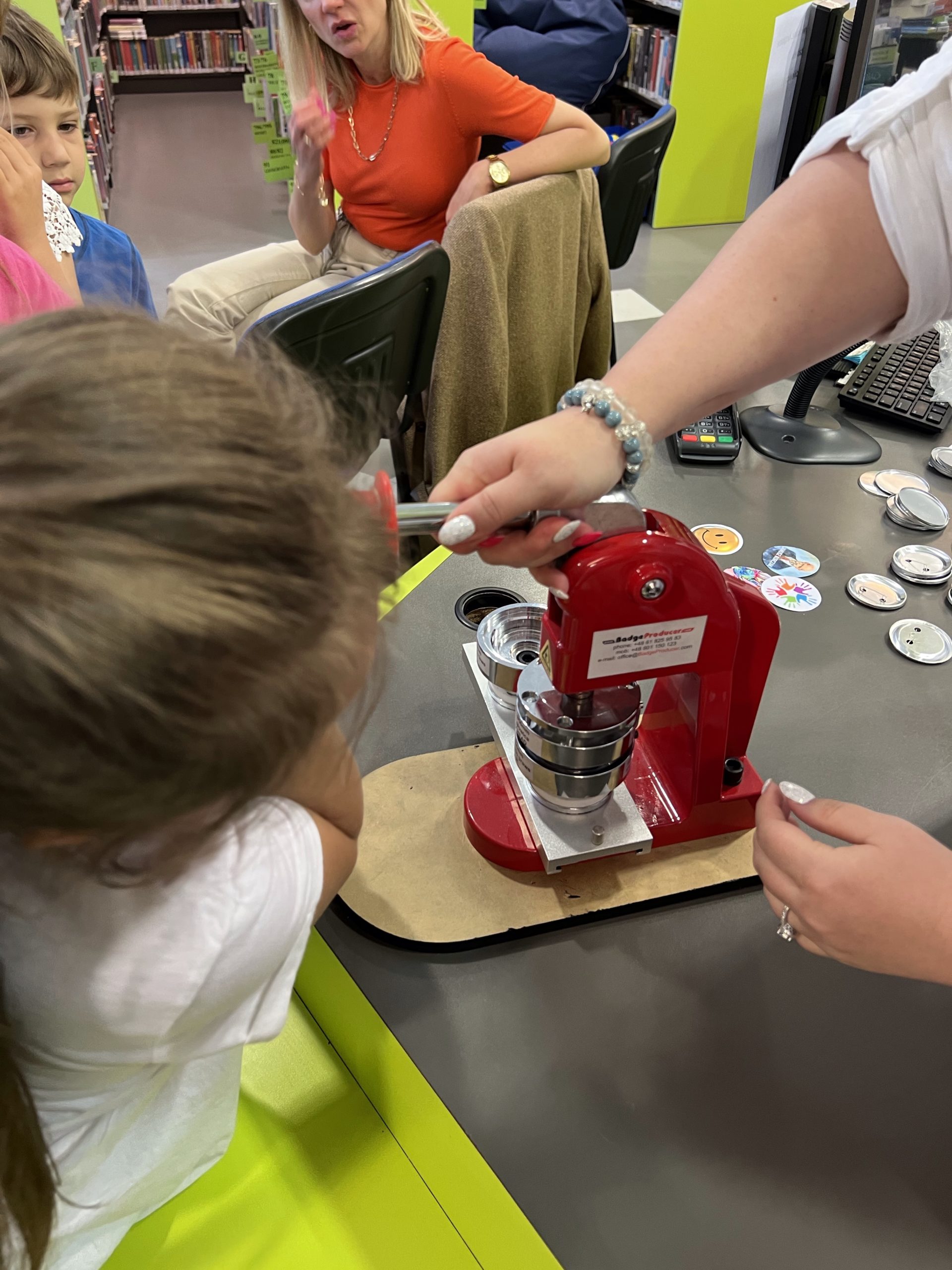 dzieci z bibliotekarzem wykonują przypinki, czerwona maszyna do robienia przypinek