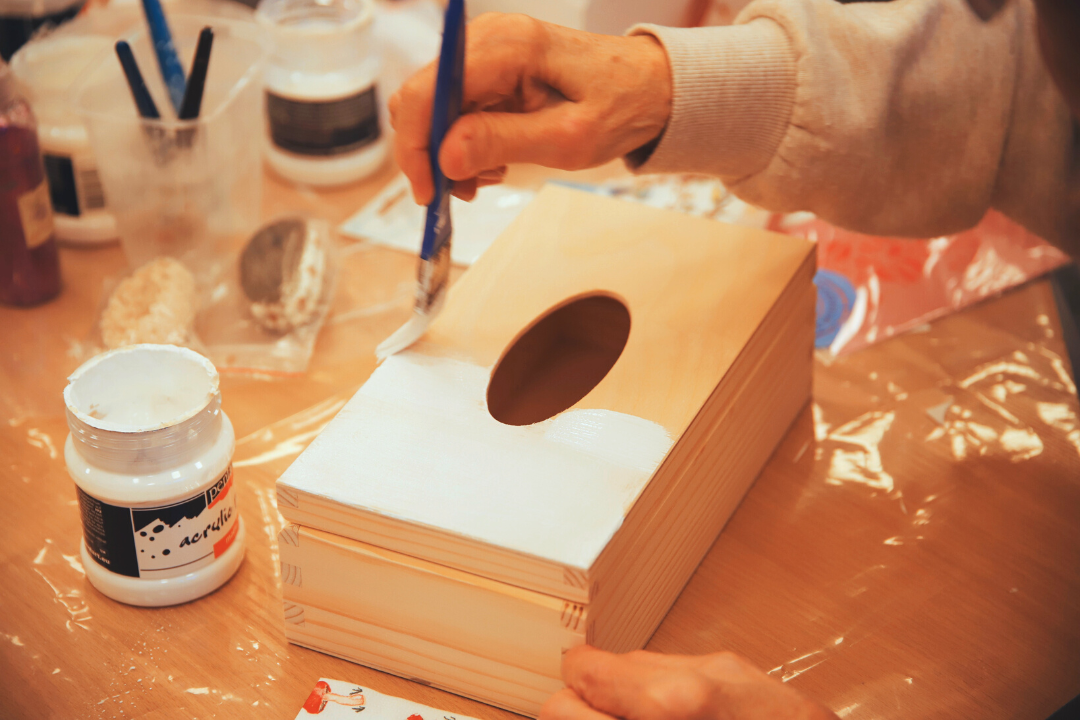 zdjęcie przedstawiające uczestniczkę warsztatów z ozdabiania breloczków metodą decoupage podczas malowania serwetnika białą farbą we wnętrzu wypożyczalni nr 16