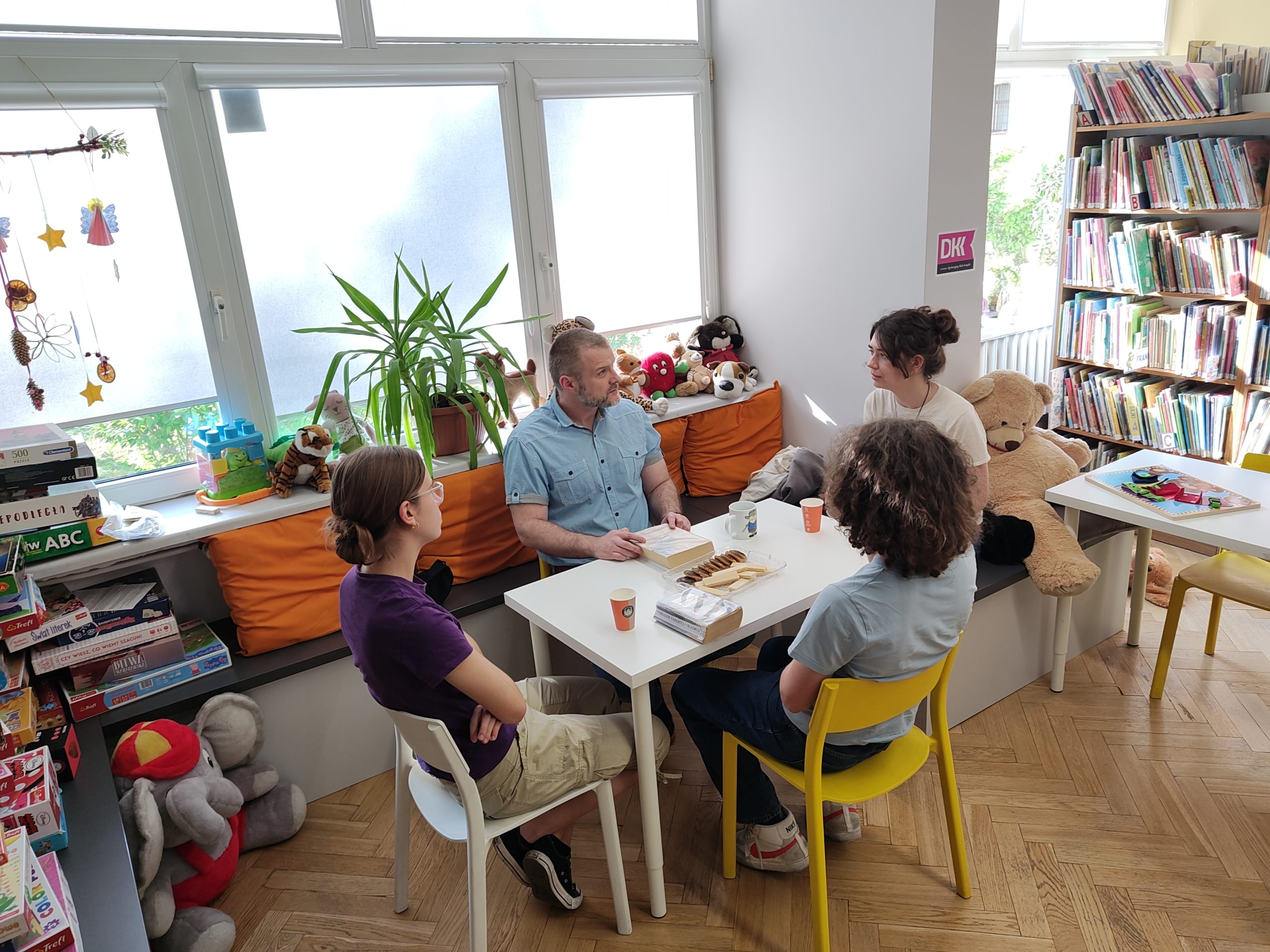grafika wyróżniająca przedstawiająca dwie dziewczyny, jednego chłopaka i bibliotekarza podczas trwania Młodzieżowego Dyskusyjnego Klubu Książki. Uczestnicy siedzą wokół białego stołu.