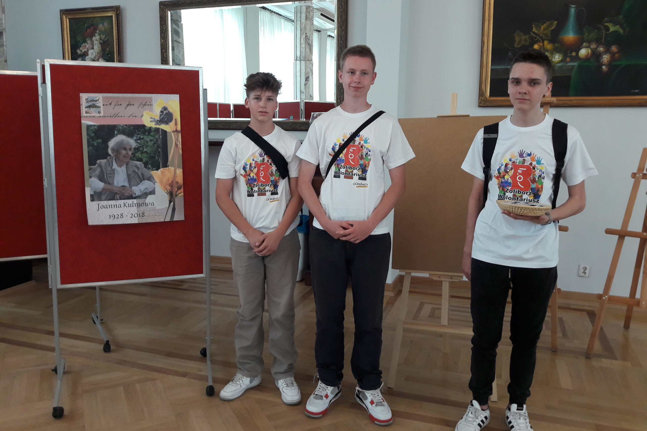 Trzech chłopców w białych koszulkach z logo wolontariatu biblioteki żoliborskiej stoi na tle wystawy. Na tablicach ekspozycyjnych wiszą kolorowe grafiki z tekstami wierszy.