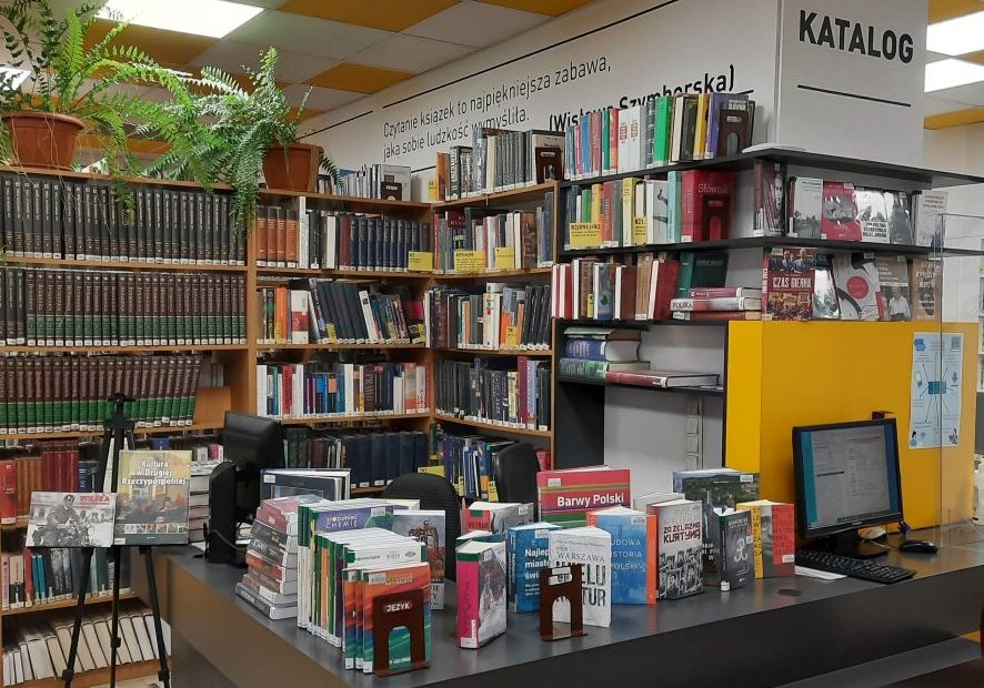 Na zdjęciu widać część sali Czytelni Naukowej nr XVI. Na ladzie bibliotecznej znajduje się wystawka publikacji ksiażkowych. W tle regały z książkami. Po prawej stronie stanowisko katalogowe.
