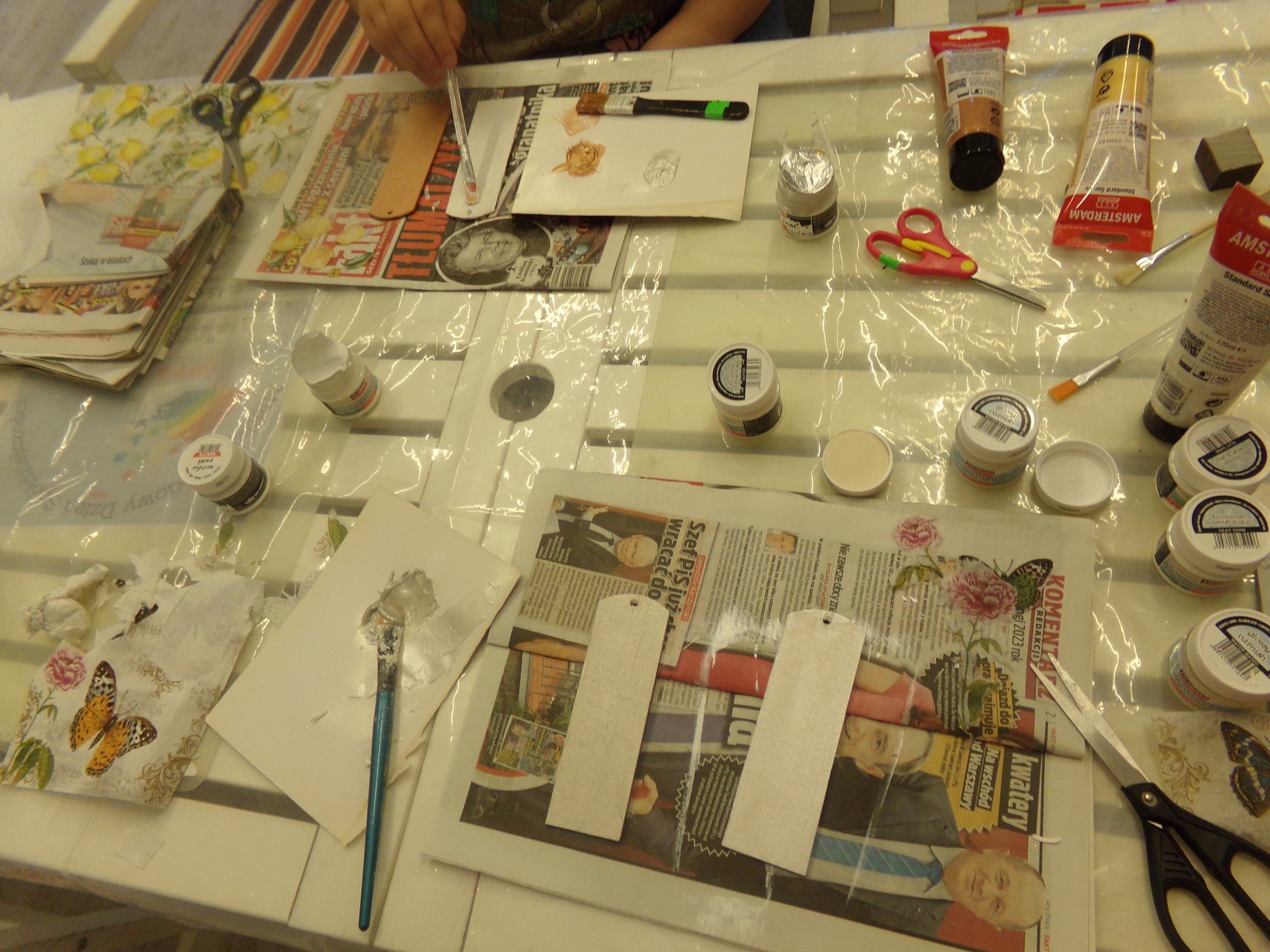 Na zdjęciu  leżą na stole materiały przygotowane do robienia zakładek metodą decoupage (folia, gazety, nożyczki, pędzle i farby).