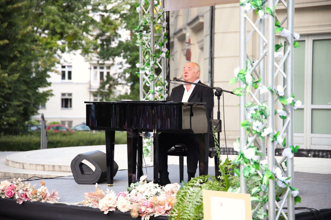 zdjęcie przedstawiające stanisława soykę grającego na fortepianie i śpiewającego podczas wydarzenia Piknik Narodowe Czytanie 2023 – „Co kto miłuje… w bibliotece znajduje” na scenie wydarzenia przed budynkiem teatru komedia w parku żywiciela przy ul. słowackiego 19a