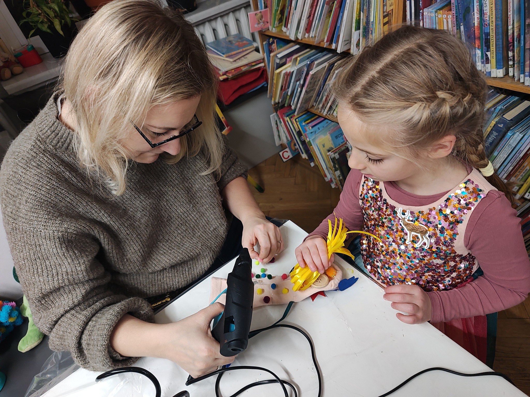 Mama pomaga córce przy wklejaniu klejem na gorąco różnych kolorowych elementów.