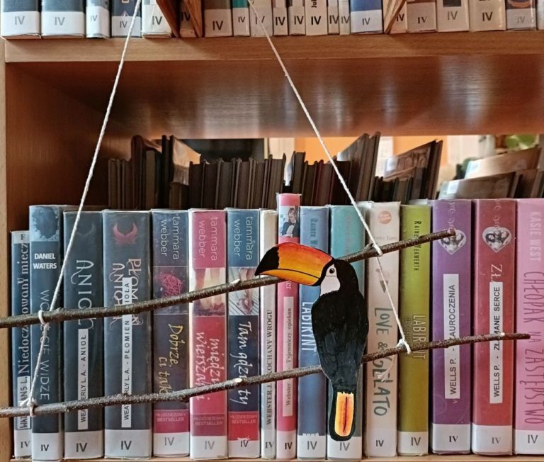 Dekoracja wisząca na regale z książkami. Konstrukcja z dwóch ułożonych równolegle do siebie patyków, związanych sznurkiem. Do jednego z patyków przyklejono papierową postać tukana.