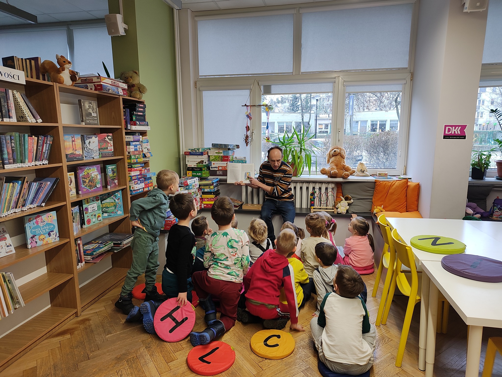 grafika wyróżniająca przedstawiająca grupę dzieci podczas słuchania czytanej przez bibliotekarza bajki.
