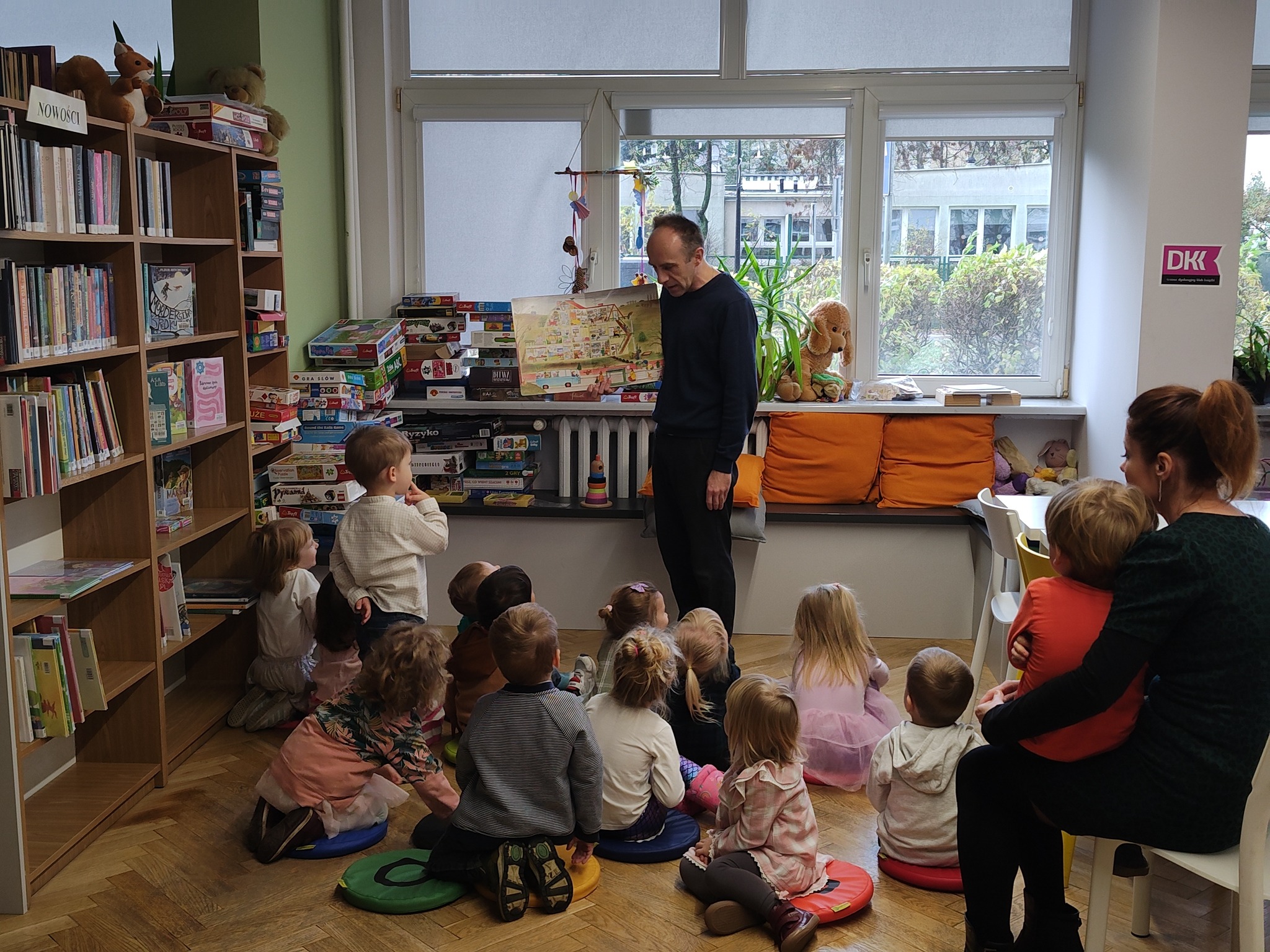 grafika wyróżniająca przedstawiająca grupę przedszkolaków, jedno dziecko po lewej stronie stoi i zadaje pytanie bibliotekarzowi.