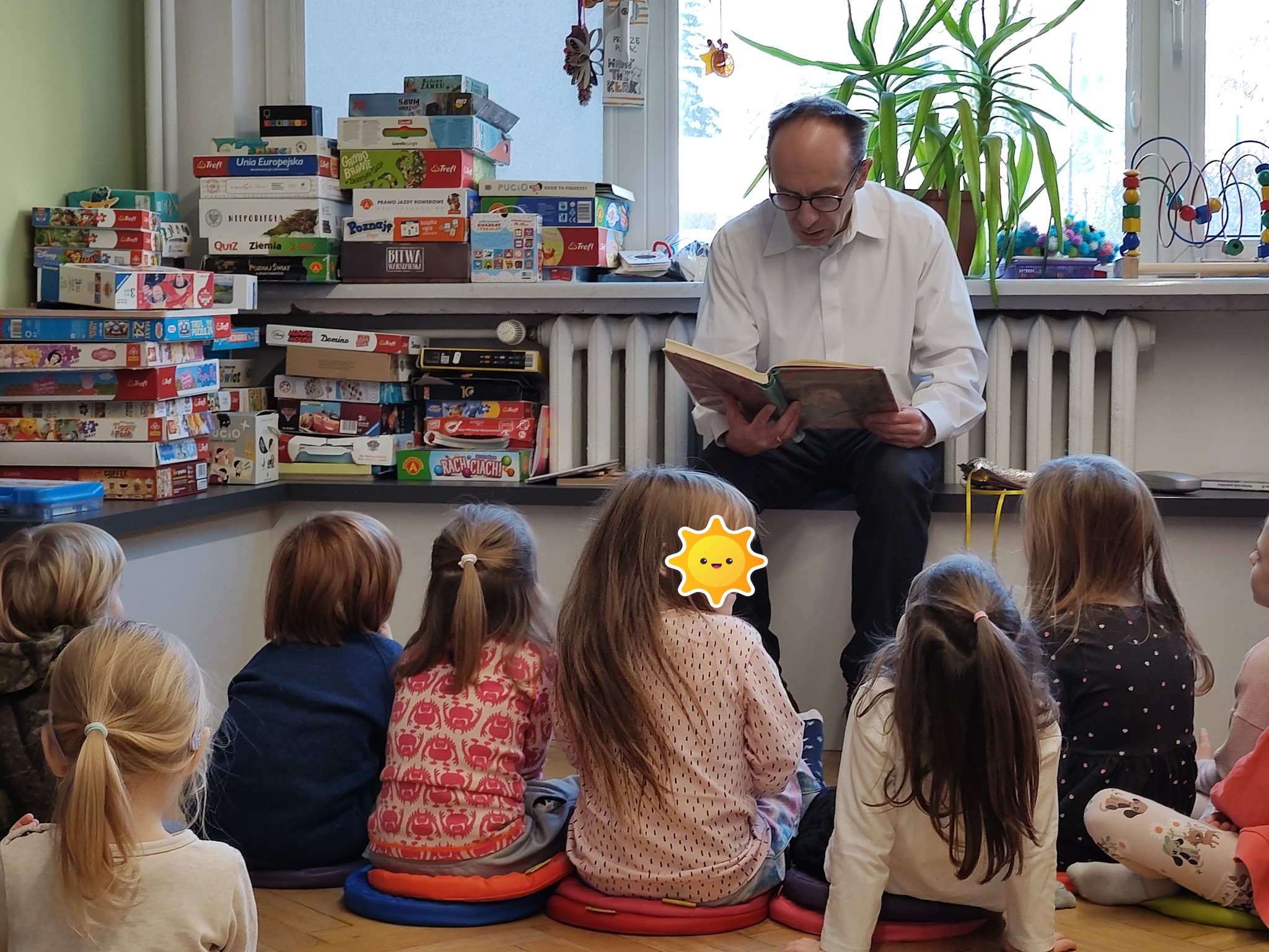 grafika wyróżniająca przedstawiająca grupkę dzieci, które słuchają czytanego przez bibliotekarza fragmentu opowiadania o "Królu Maciusiu Pierwszym" Janusza Korczaka. Bibliotekarz jest przebrany za Janusza Korczaka - ma białą koszulę, okulary i ciemne spodnie.
