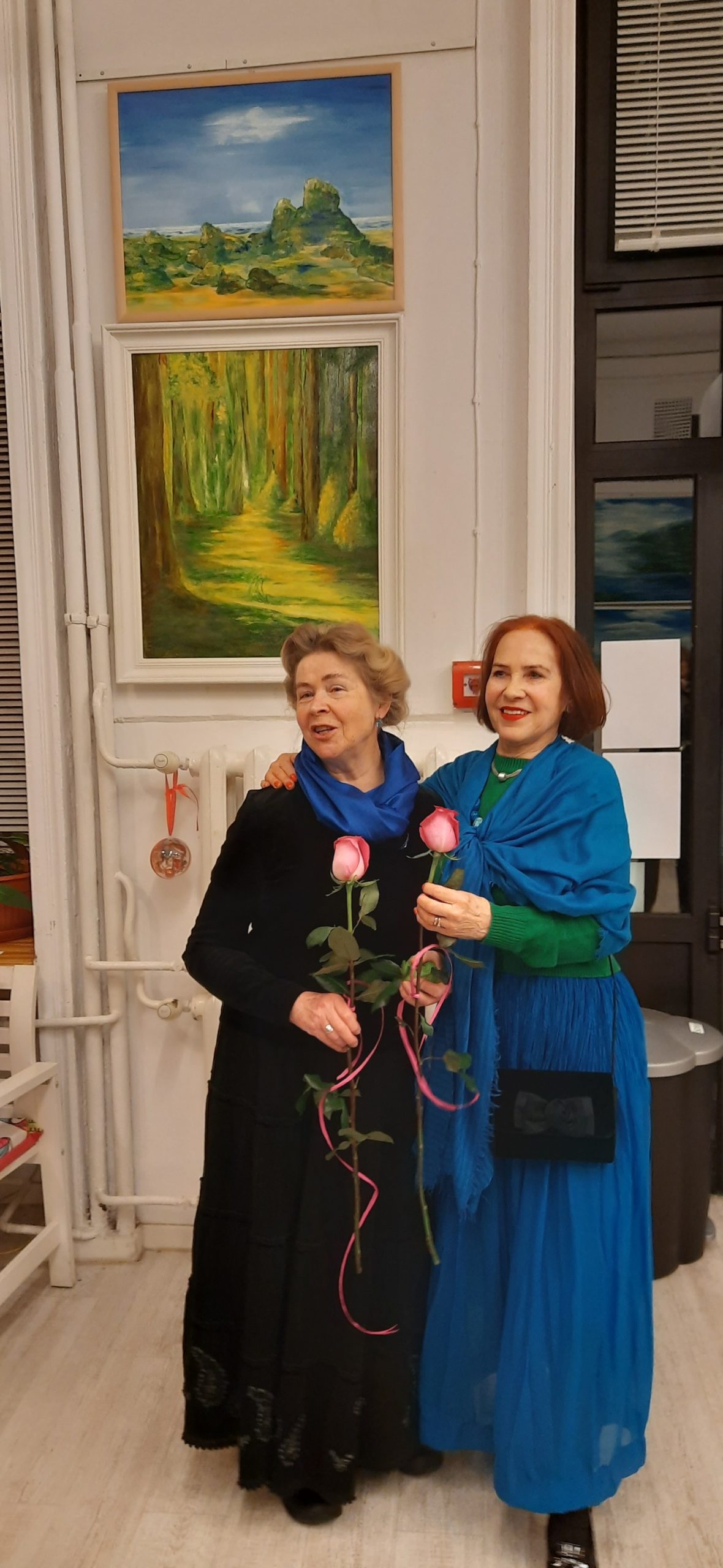 Zdjęcie przedstawia dwie uśmiechnięte kobiety, każda z kwiatem róży. W tle na białej ścianie wiszą obrazy olejne z pejzażami.