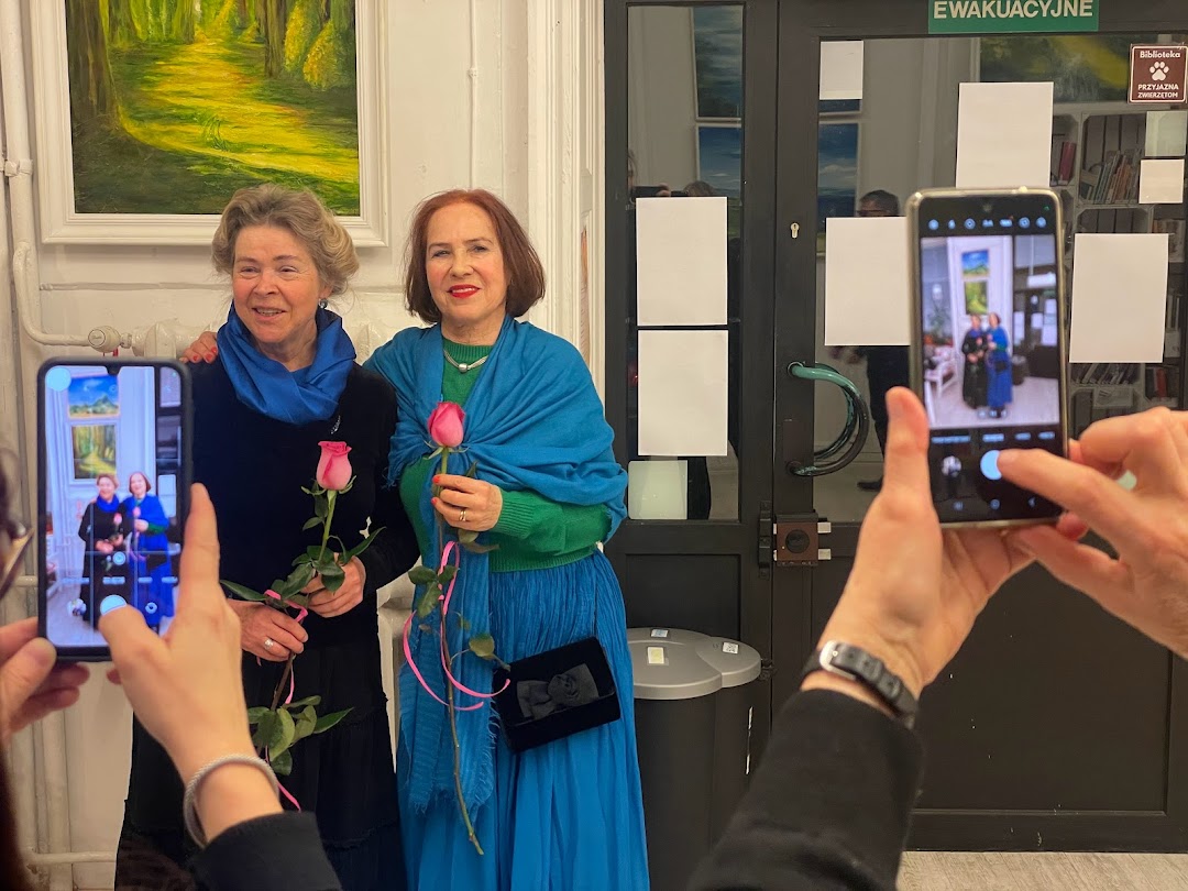 Zdjęcie przedstawia dwie uśmiechnięte kobiety pozujące do zdjęć, każda z kwiatem róży. W tle na białej ścianie wisi obraz olejny z pejzażem. Na pierwszym planie widać ręce osób robiących zdjęcie telefonami.