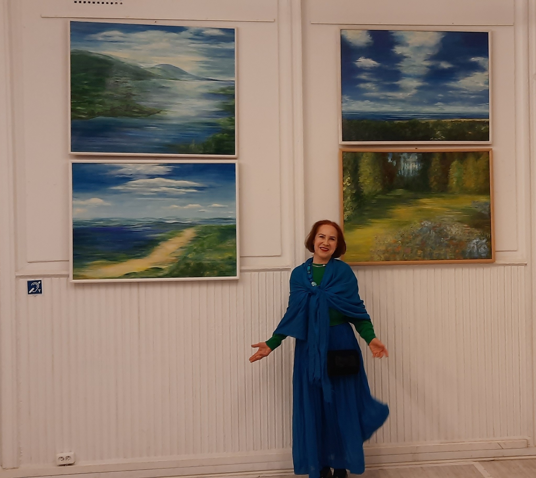 Zdjęcie przedstawia uśmiechniętą kobietę rozkładającą szroko ręce. W tle na białej ścianie wiszą cztery obrazy olejne z pejzażami.