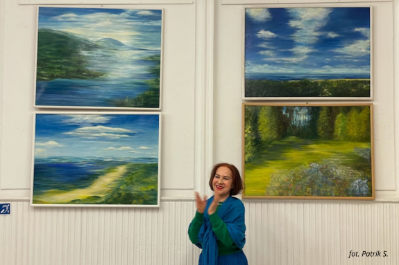 Zdjęcie przedstawia uśmiechniętą kobietę składającą ręce do oklasków. W tle na białej ścianie wiszą cztery obrazy olejne z pejzażami.