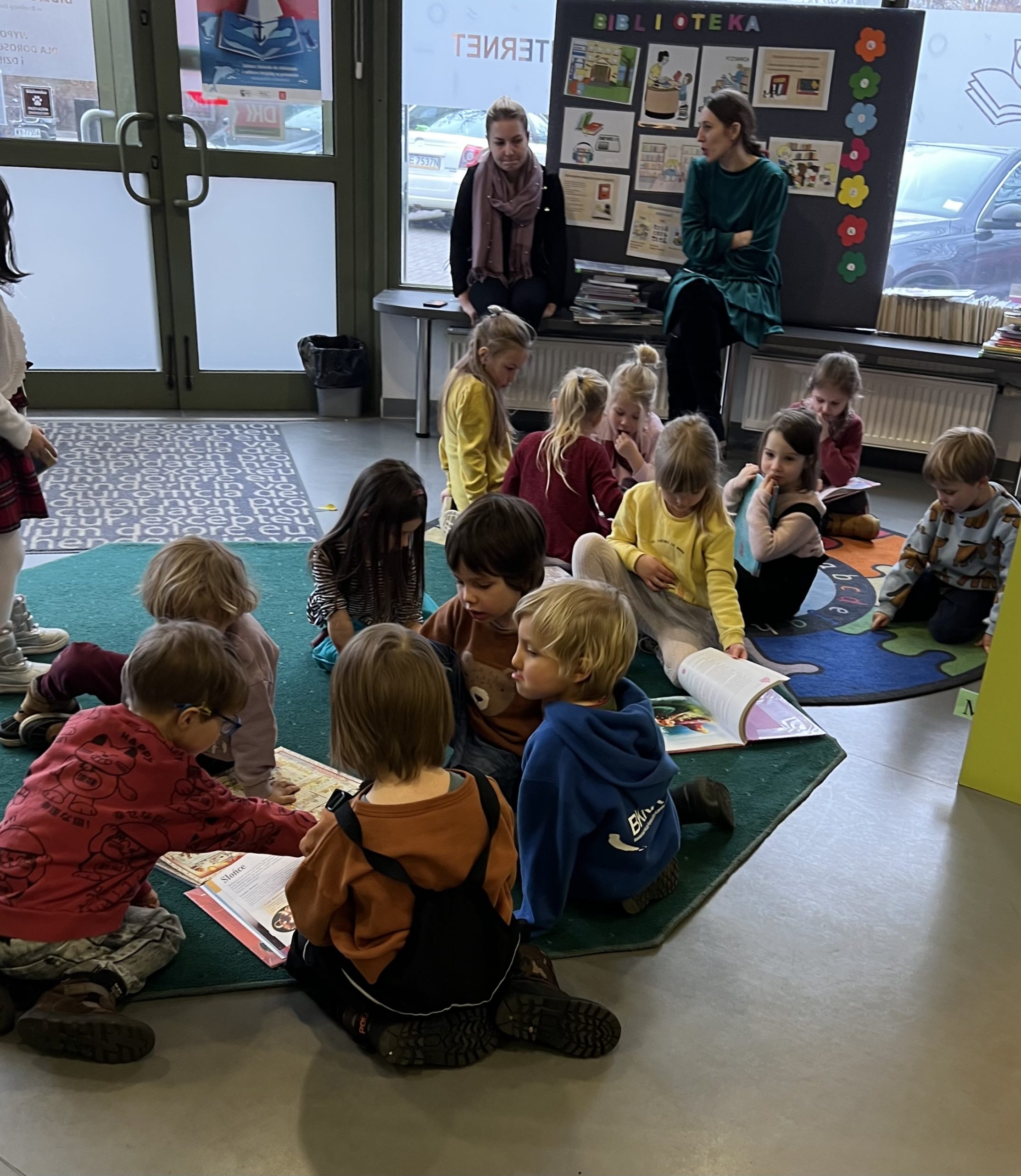 grupa dzieci siedzi na podłodze i ogląda książki, w tle dwie nauczycielki siedzą przy tablicy, na której wiszą kolorowe plansze