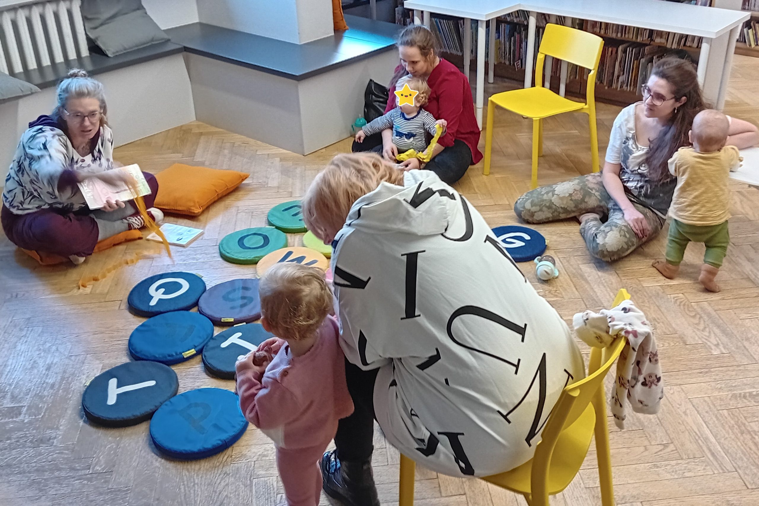 grafika wyróżniająca przedstawiająca rodziców z dziećmi podczas zabawy z książką i kolorową krepiną. Bibliotekarka trzyma w ręku książkę i krepinę a mamy z dziećmi słuchają opowiadania czynnie uczestnicząc w zabawie.