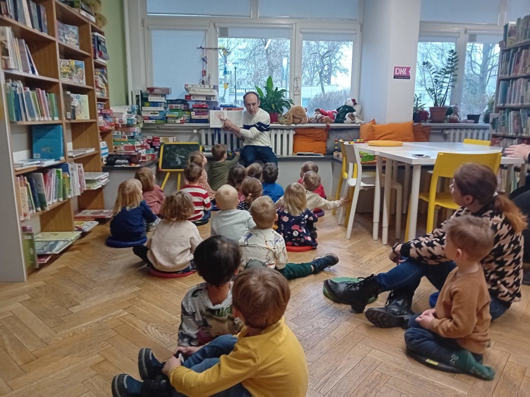 grafika wyróżniająca przedstawiająca dzieci, które słuchają czytanej książki i oglądają ilustracje pokazywane przez bibliotekarza.