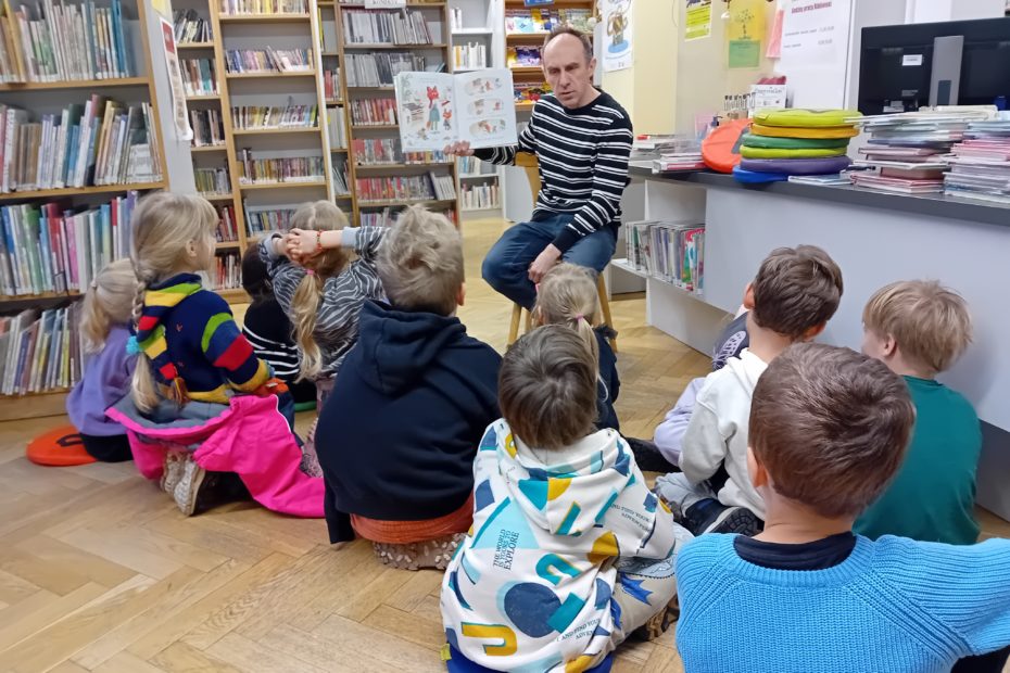 grafika wyróżniająca przedstawiająca w sali bibliotecznej dzieci siedzące na poduszkach i słuchające opowiadania czytanego przez bibliotekarza. Bibliotekarz pokazuje co znajduje się w książce.