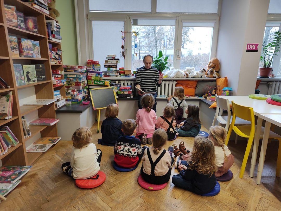 grafika wyróżniająca przedstawiająca dzieci słuchających bibliotekarza w trakcie czytania książki.
