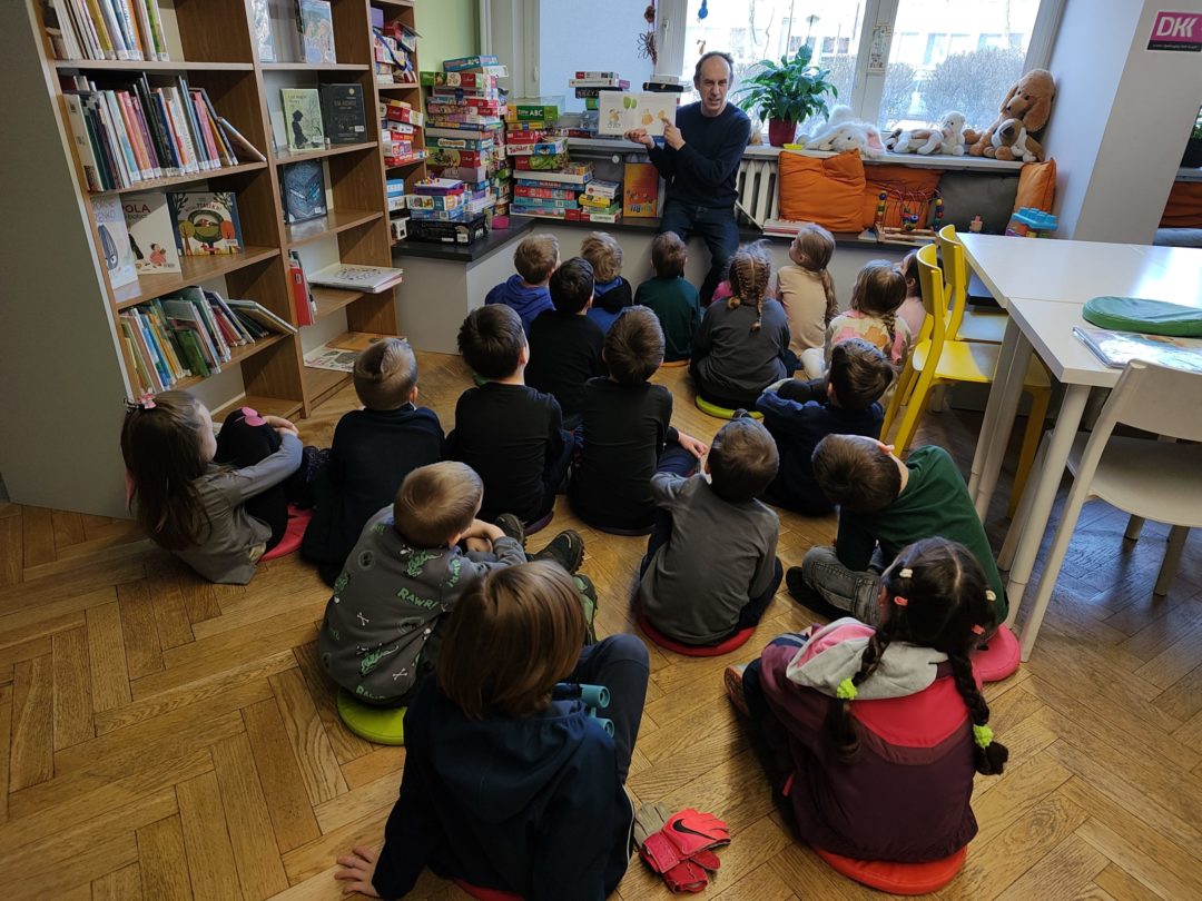 grafika wyróżniająca przedstawiająca grupę dzieci podczas czytania bajek. Bibliotekarz pokazuje im ilustracje.