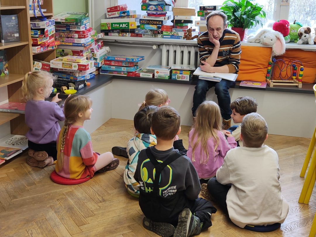 grafika wyróżniająca przedstawiająca grupę dzieci podczas rozmowy o czytanym opowiadaniu. Bibliotekarz słucha ich wypowiedzi podpierając ręką głowę.