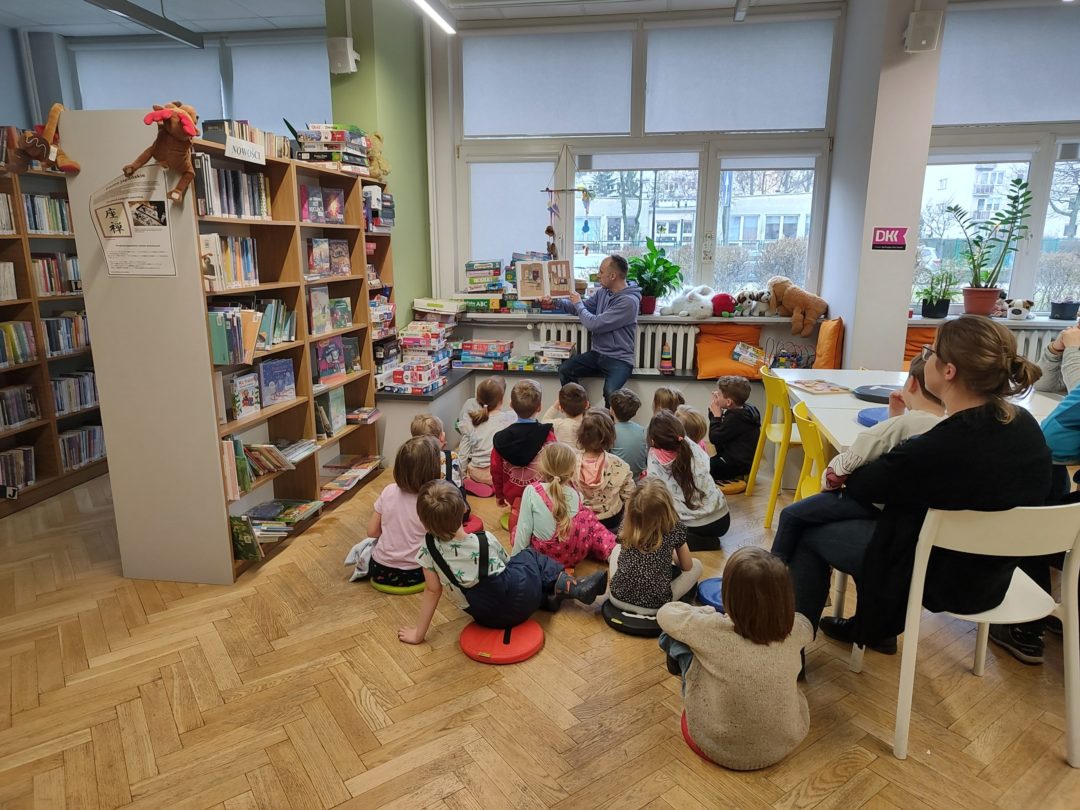 grafika wyróżniająca przedstawiająca grupę dzieci patrzących na obrazki książki , którą pokazuje bibliotekarz.