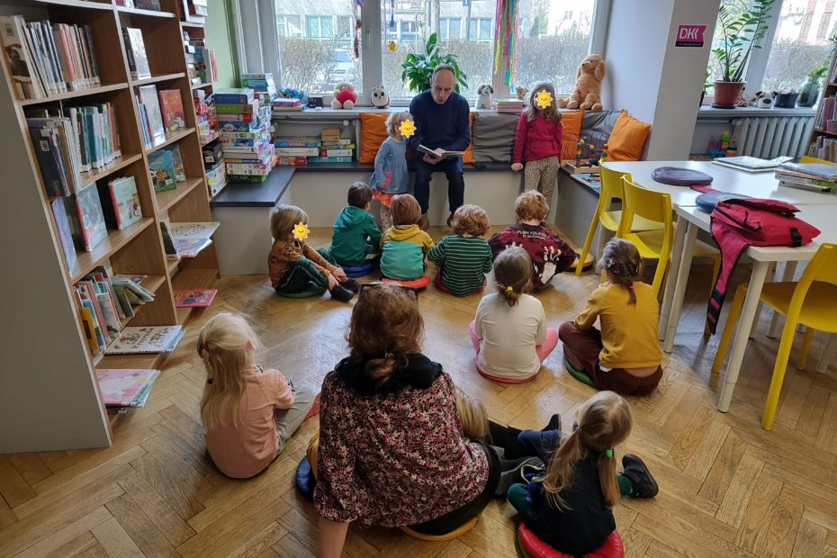 grafika wyróżniająca przedstawiająca grupę dzieci podczas czytania bajek. Bibliotekarz czyta książkę a po jego lewej i prawej stronie stoją dziewczynki.