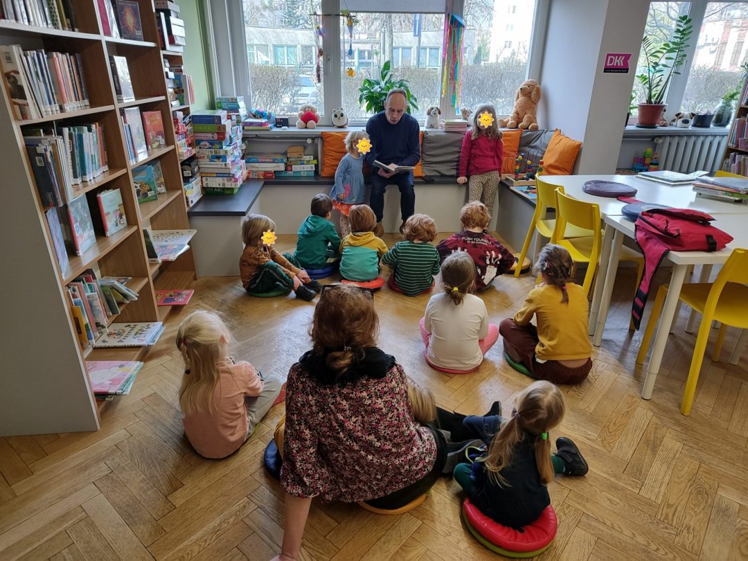 grafika wyróżniająca przedstawiająca grupę dzieci podczas czytania bajek. Bibliotekarz czyta książkę a po jego lewej i prawej stronie stoją dziewczynki.