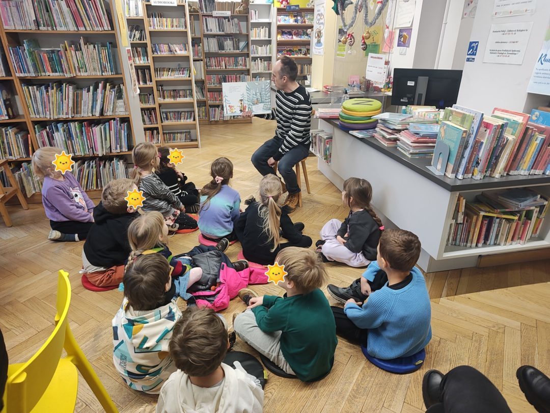 grafika wyróżniająca przedstawiająca w sali bibliotecznej dzieci siedzące na poduszkach i słuchające opowiadania czytanego przez bibliotekarza. Bibliotekarz pokazuje co znajduje się w książce.