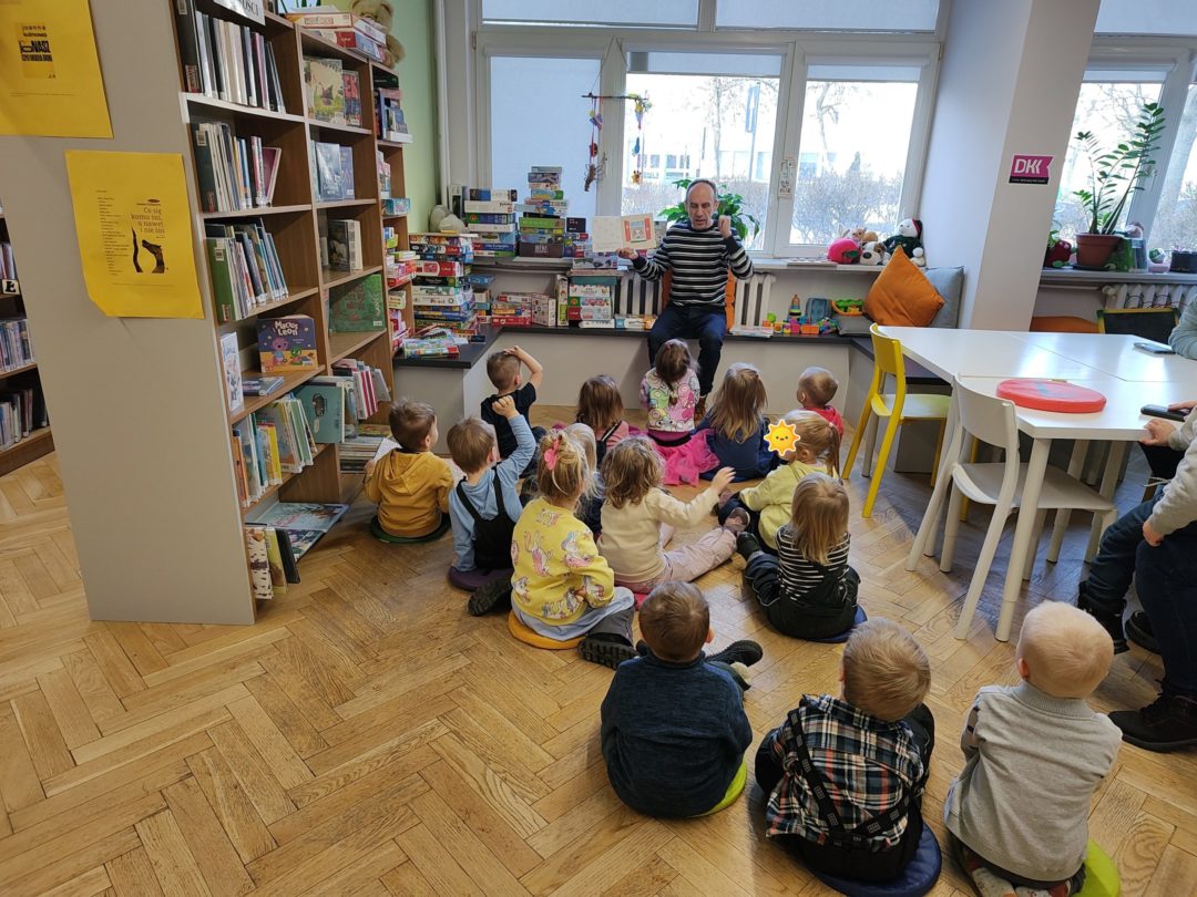 grafika wyróżniająca przedstawiająca w sali bibliotecznej dzieci siedzące na poduszkach i słuchające opowiadania czytanego przez bibliotekarza. Bibliotekarz pokazuje co znajduje się w książce.