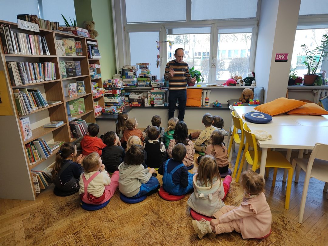 grafika wyróżniająca przedstawiająca grupę dzieci, które znajdują się w sali bibliotecznej i słuchają bibliotekarza, który opowiada im jak mogą korzystać z przestrzeni biblioteki i książek.