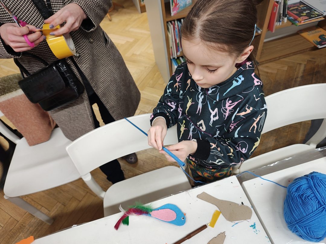 grafika wyróżniająca przedstawiająca dziewczynkę podczas warsztatów. Wykonuje ptaka z piankowego arkusza w kolorze niebieskim, który ma z tego samego materiału różowe skrzydełka. Ogon zrobiony jest z kolorowych piórek.