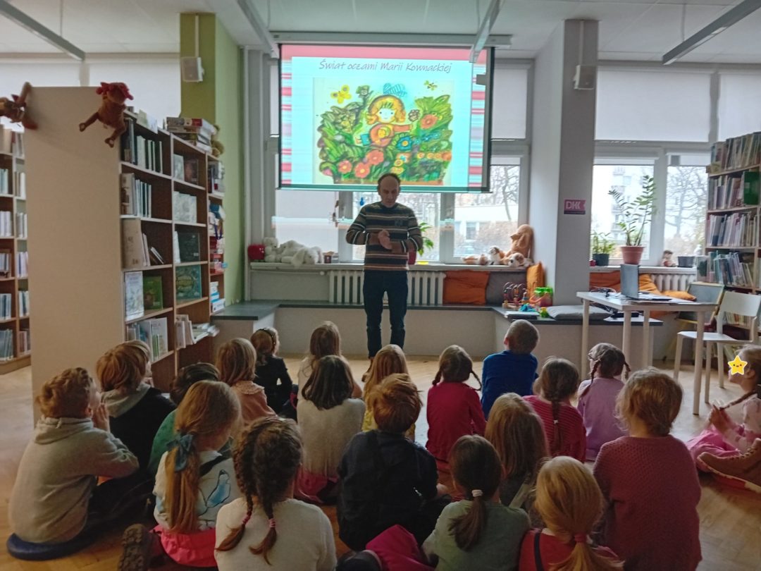 grafika wyróżniająca przedstawiająca uczestników podczas spotkania o Marii Kownackiej. Dzieci słuchają bibliotekarza, który ma za sobą ekran, na którym widnieje slajd z ilustracją jednej z książek Marii Kownackiej.