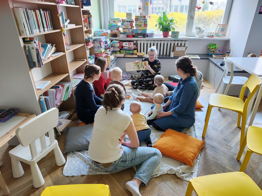 grafika wyróżniająca przedstawiająca prowadzącą zajęcia podczas czytania wiersza "Ptasie Radio" Juliana Tuwima dla grupy dzieci i ich mam.