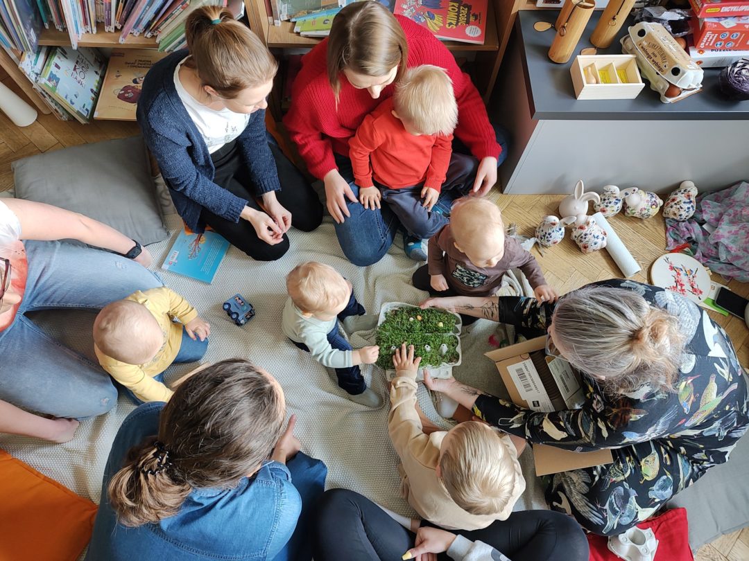 grafika wyróżniająca przedstawiająca grupę mam z dziećmi, które oglądają razem z prowadzącą rzeżuchę wysianą w papierowym pudełku po jajkach.