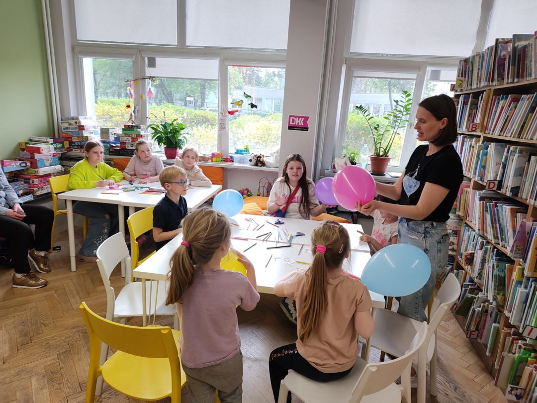 grafika wyróżniająca przedstawiająca uczestników podczas pierwszego eksperymentu z elektrostatyki. Dzieci trzymają w dłoniach kolorowe baloniki a prowadząca warsztaty opowiada jak ma wyglądać eksperyment.