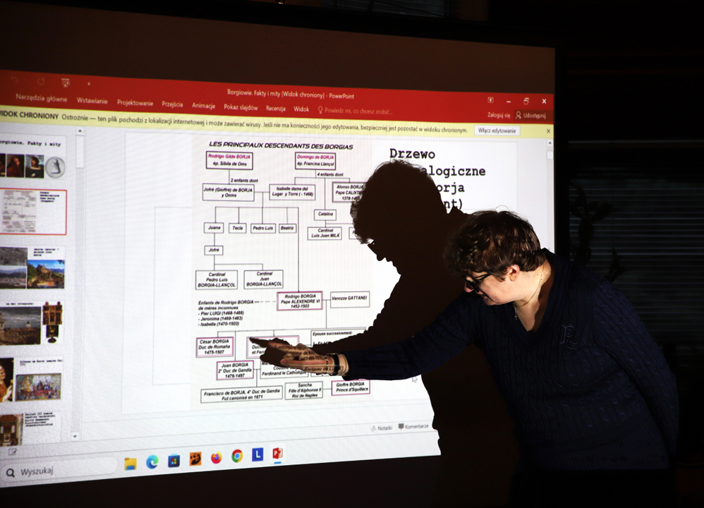 Zdjęcie szóstej prelekcji z cyklu wykładów o sztuce i historii: "Borgiowie - fakty i mity" przedstawiające Panią Magdalenę Tworzowską pokazującą palcem na slajdzie na rzutniku z napisami Drzewo genealogiczne rodziny Borja we wnętrzu Wypożyczalni nr 16
