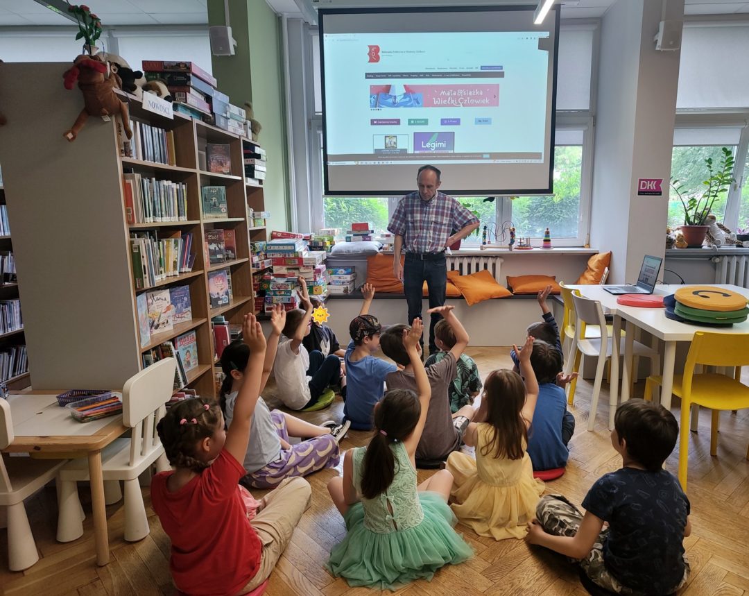 grafika wyróżniająca przedstawiająca grupę uczniów klasy pierwszej podczas lekcji bibliotecznej, którą prowadzi bibliotekarz