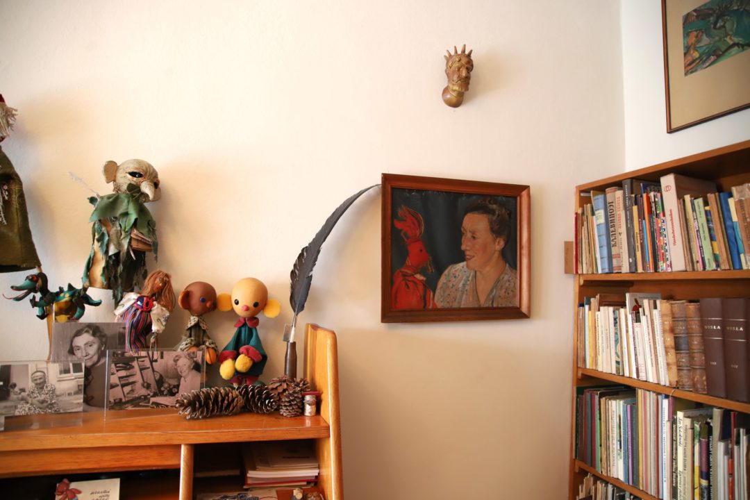 zdjęcie z wnętrza Muzeum Książki Dziecięcej lub inaczej Izby Pamięci Marii Kownackiej, która mieści się na warszawskim Żoliborzu przy ulicy Słowackiego 5/13 m. 74. zdjęcie ściany z półką ze zdjęciami, figurkami plastusiów oraz regału z książkami i malowanym portretem marii kownackiej