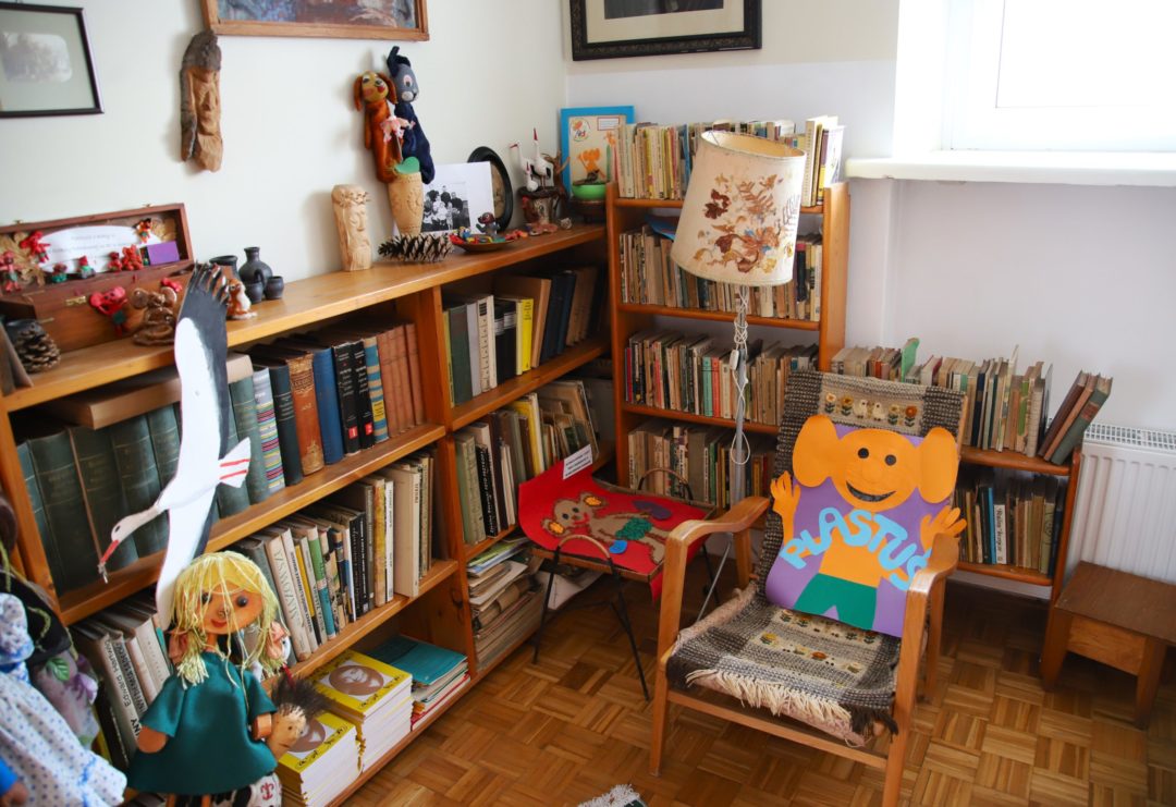 zdjęcie z wnętrza Muzeum Książki Dziecięcej lub inaczej Izby Pamięci Marii Kownackiej, która mieści się na warszawskim Żoliborzu przy ulicy Słowackiego 5/13 m. 74. zdjęcie rogu z regałami z książkami, fotelem z plakatem przedstawiającym plastusia oraz różnymi przedmiotami: figurkami, lalkami, lampą, stołkiem