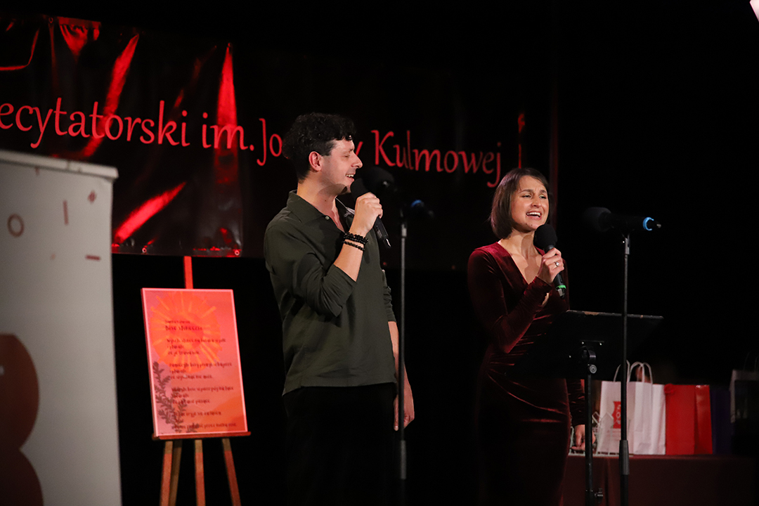 Zdjęcie z II Żoliborskiego Konkursu Recytatorskiego im. Joanny Kulmowej zorganizowanego przez Bibliotekę Publiczną w Dzielnicy Żoliborz, Społeczny Dom Kultury WSM oraz Fundację Joanny i Jana Kulmów. Artyści występujący na scenie Społecznego Domu Kultury śpiewają przy mikrofonach. W tle baner z napisem Żoliborski Konkurs Recytatorski im. Joanny Kulmowej oraz sztalugi z fragmentami utworów Joanny Kulmowej.